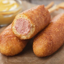 3 Corn Dogs on a brown napkin with a bowl of mustard in the background. One of the corndogs is half eaten.