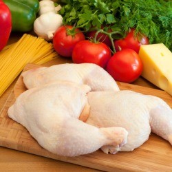 Three raw chicken leg quarters piled on cutting board in front of tomato, lettuce, peppers ,and a wedge of parmesan cheese