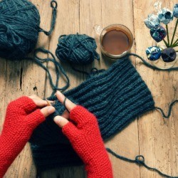 Person wearing red fingerless gloves knitting something grey.